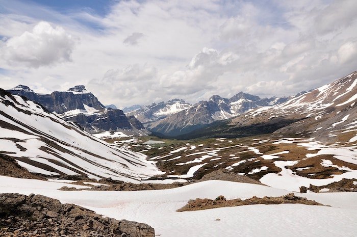 Una foto paisajística de montañas, nubes, cielo azul y un suelo rocoso y estéril con nieve.