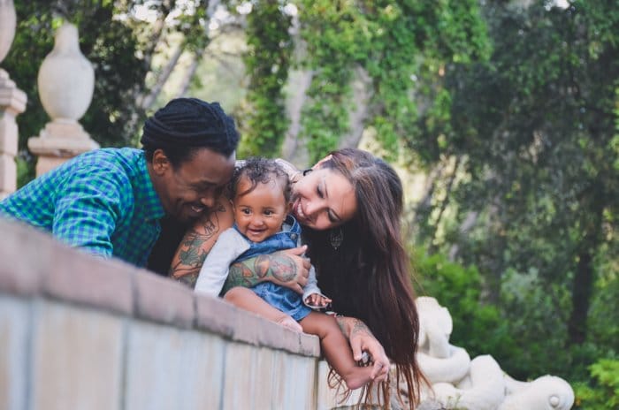 Foto de retrato de familia de una pareja y un bebé pequeño apoyado en una pared. 
