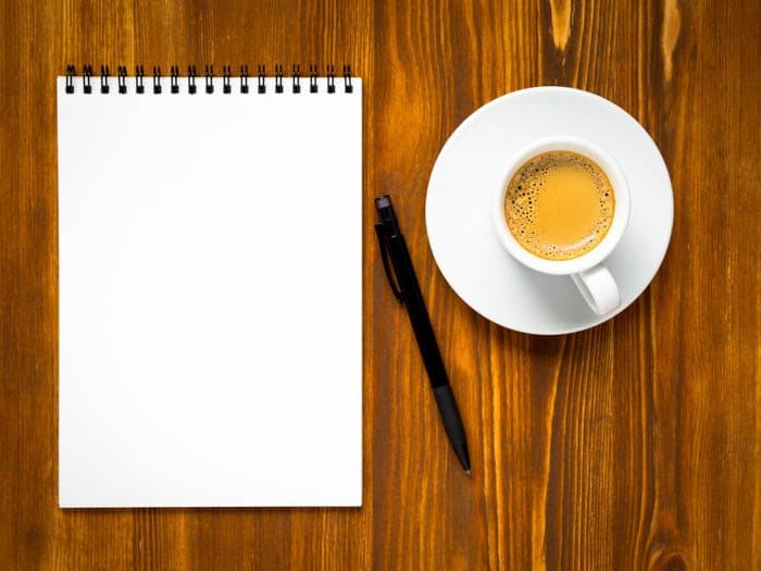 Bloc de notas abierto con una página en blanco para escribir una lista de tareas pendientes de viaje, una taza de café y un bolígrafo en la mesa marrón de madera