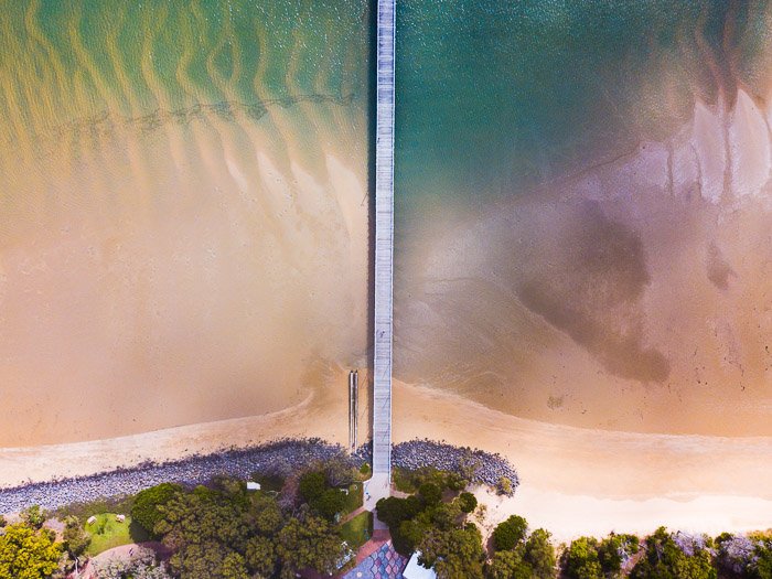 Fotografía de drones hacia abajo de un largo puente de madera en aguas poco profundas que captura la simetría combinando características naturales y creadas por el hombre.