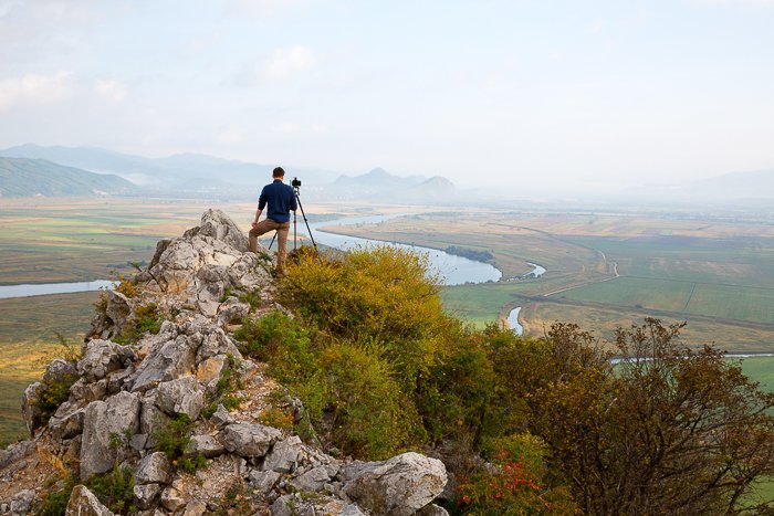 un fotógrafo se encuentra en lo alto de una colina rocosa con vistas a un paisaje verde y al río