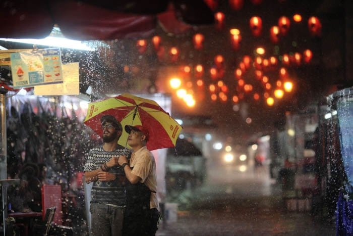 Dos hombres en un mercado al aire libre lluvioso por la noche, iluminados por farolillos coloridos