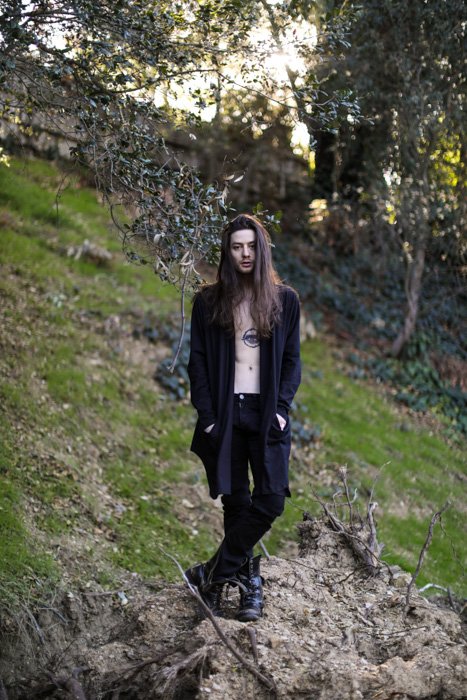 Retrato de un hombre de pelo largo, ropa negra y pecho tatuado, de pie en una zona forestal, mirando a la cámara.  Mejore sus habilidades fotográficas hoy.