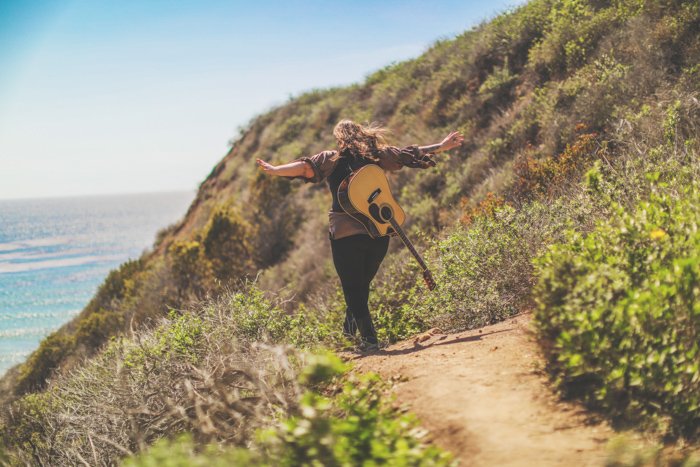 Foto al aire libre de una persona con la guitarra acústica en la espalda, alejándose de la cámara por un camino costero, con los brazos extendidos.  Mejore sus habilidades fotográficas hoy.