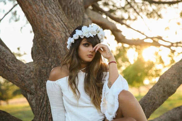 Luminoso y aireado retrato de una niña con vestido blanco con flores blancas en el pelo, sentada frente a un árbol.  Mejore sus habilidades fotográficas hoy