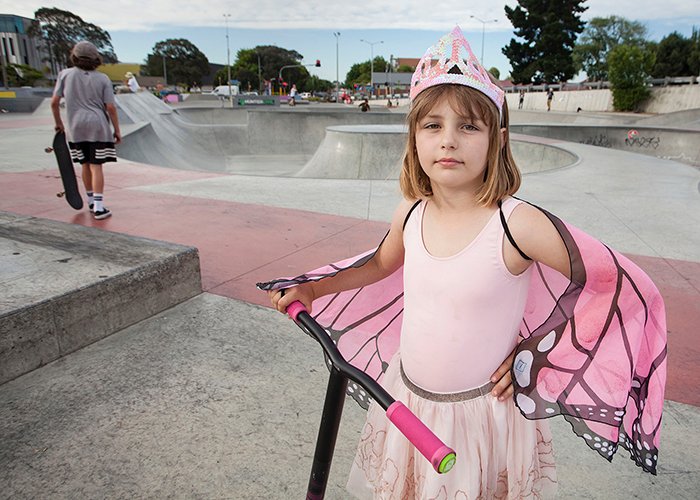 Niña en traje de mariposa princesa rosa con un scooter, parque de patinaje en el fondo