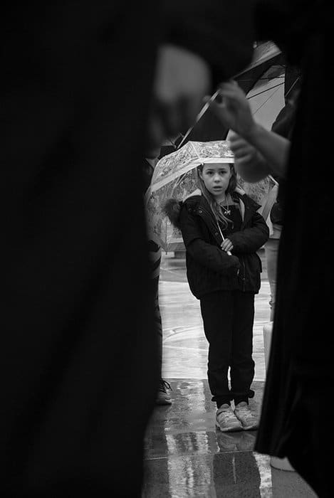 Fotografía documental en blanco y negro de una niña sosteniendo un paraguas enmarcado por figuras más grandes.