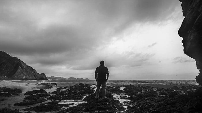 Fotografía costera monocroma de un hombre de pie sobre las rocas mirando hacia el océano con una relación de aspecto de 16: 9