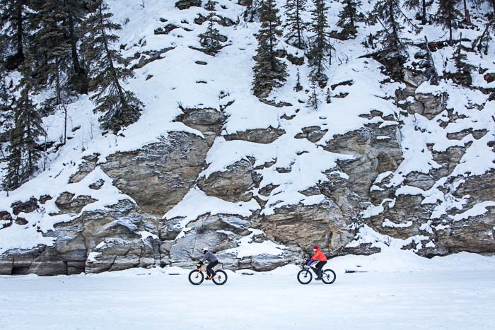 Vista desde lejos de dos ciclistas en la nieve.  La ladera de la montaña está al fondo.