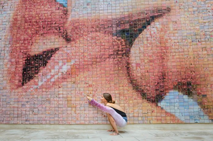 Fotografía de yoga artístico de un modelo en pose de luciérnaga frente a una pared de mosaico que muestra a dos personas besándose