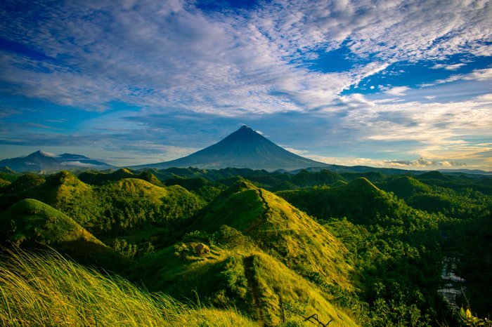 una imagen de una montaña en el fondo con colinas verdes en el primer plano y el cielo azul y las nubes arriba