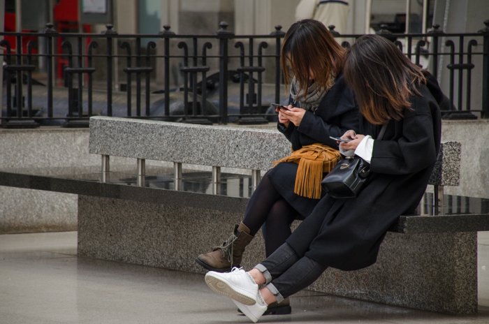 Foto de la calle de dos niñas sentadas en un banco y usando teléfonos inteligentes, mostrando tonos medios y oscuros en imágenes