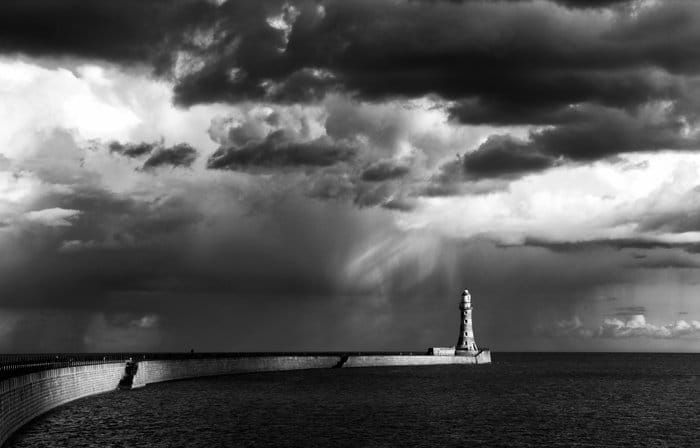 Una impresionante foto en blanco y negro del faro bañado por el sol en un promontorio con nubes oscuras sobre su cabeza