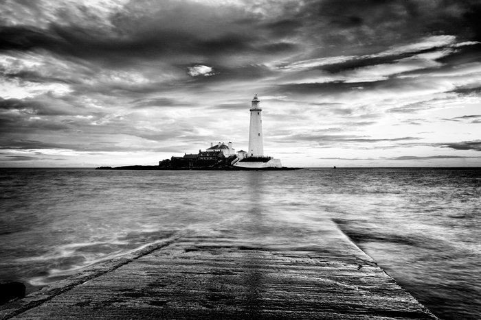 Vista costera en blanco y negro a través del agua mirando hacia un faro, rodada con una larga exposición