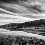 Una fotografía en blanco y negro de un cielo rayado de nubes reflejado en el agua con la colina en segundo plano.