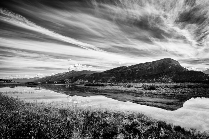 Una fotografía en blanco y negro de un cielo rayado de nubes reflejado en el agua con la colina en segundo plano.