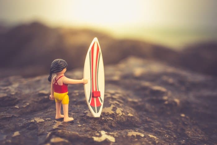 Un personaje de playmobil con una tabla de surf posada en una playa rocosa, ideas creativas para fotografías de playa.