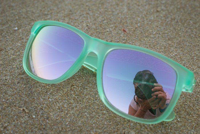Un par de gafas de sol con montura verde descansando sobre la arena con el reflejo de los fotógrafos en un vaso, ideas geniales para fotografías de playa. 