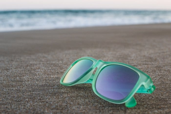 Un par de gafas de sol con montura verde descansando sobre la arena, las olas del mar borrosas en el fondo