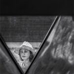 Fotografía en blanco y negro de una niña con sombrero blanco enmarcada por detalles arquitectónicos.  Fotografía callejera creativa