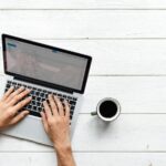 Foto de las manos de un hombre escribiendo en una computadora portátil con una taza de café al lado