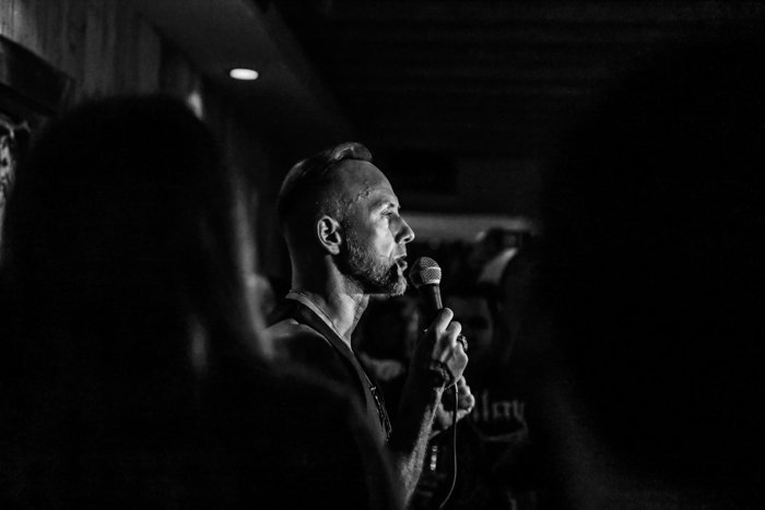 Fotografía de un evento en blanco y negro de un hombre hablando o cantando en un micrófono en el escenario