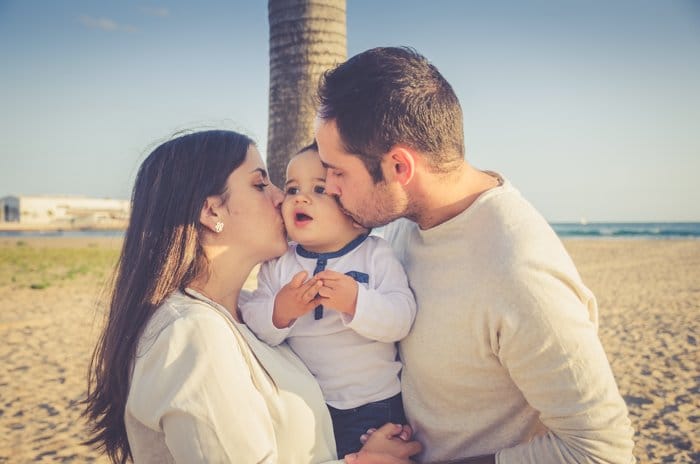 Dulce retrato de un hombre y una mujer sosteniendo a un bebé pequeño y besando sus mejillas en una playa - composición de retratos familiares