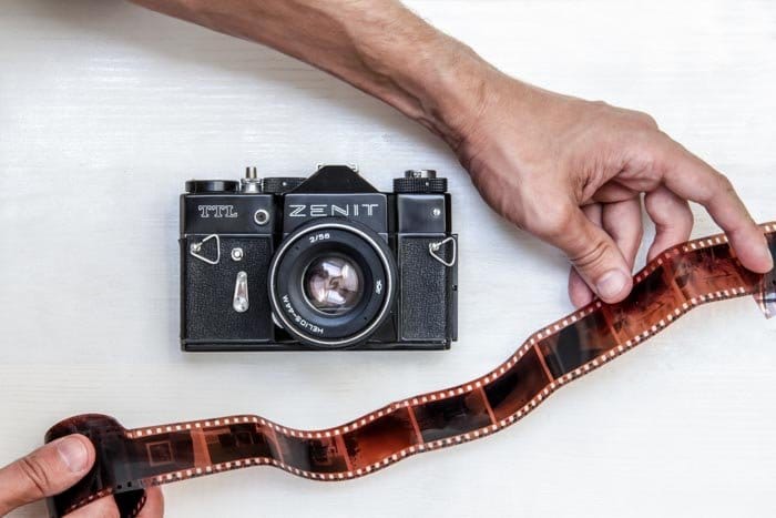 Una fotografía cenital de una cámara de cine Zenit sobre fondo blanco con una persona sosteniendo un rollo de película negativa al lado - usando película para fotografía callejera 