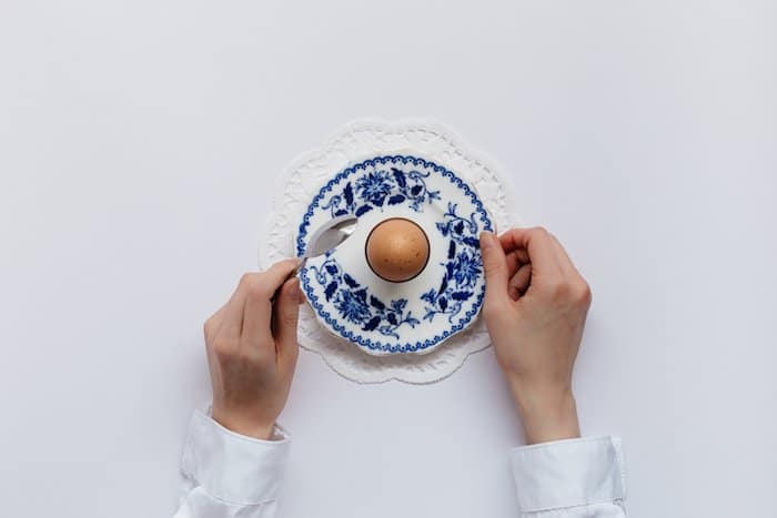 fotografía plana de comida: Manos sosteniendo cubiertos sobre un solo huevo cocido en un plato estampado sobre un fondo completamente blanco