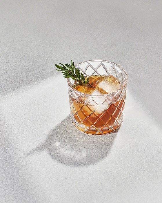 Una bebida alcohólica con hielo y romero en un vaso de vidrio utilizado como accesorios de fotografía de alimentos