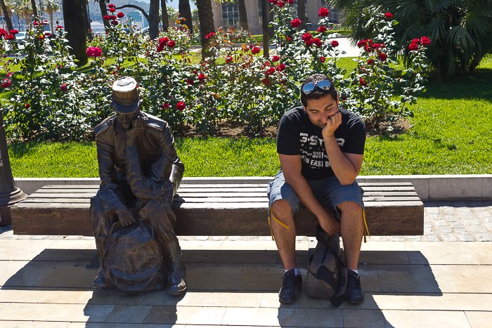 al aire libre, frente a un ramo de flores rojas, un hombre se sienta junto a una estatua, imitando su postura de pensamiento