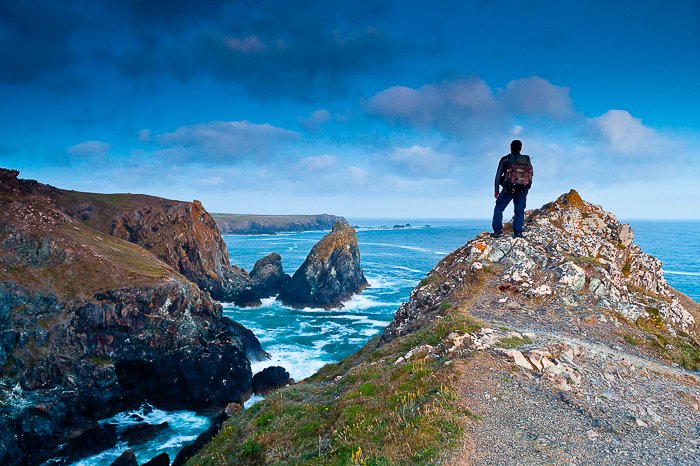 Vista desde atrás de un hombre parado en la cima de un afloramiento de roca con vista a una costa rocosa y un mar y cielo azul brillante