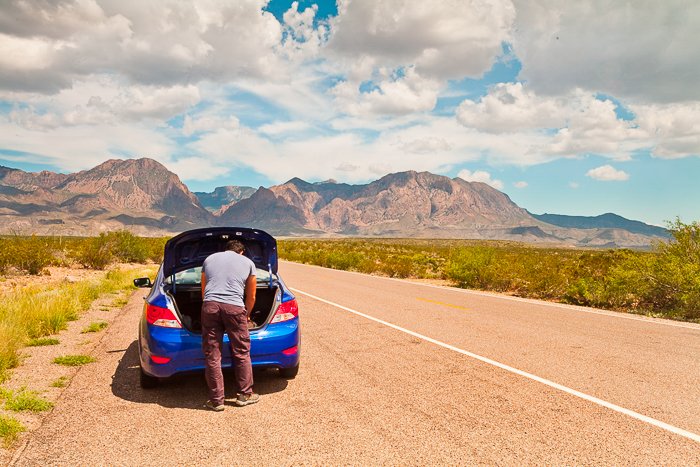 Hombre mirando el maletero de su coche azul, detenido al lado de un camino marrón pálido que conduce a las montañas en la distancia
