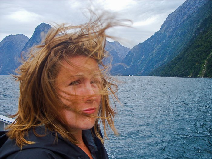 Una mujer tomándose una selfie frente a un lago azul, cielo y montañas, el viento soplando su corto cabello rubio en su rostro
