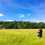 hombre con una cámara caminando por un campo de hierba verde brillante bajo un cielo azul profundo