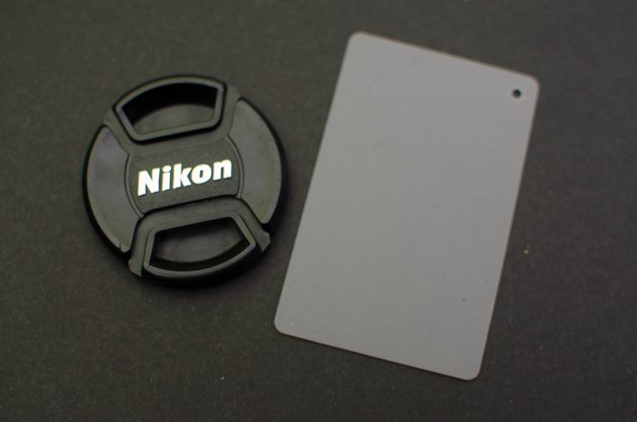 Una tapa de lente Nikon sobre fondo gris con una tarjeta gris de fotografía al lado