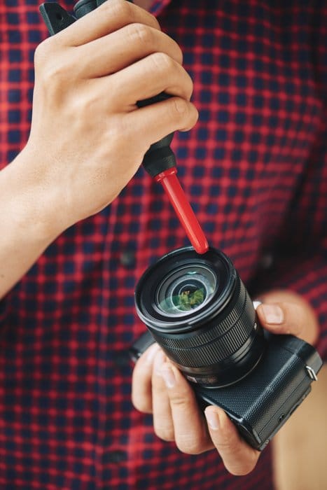 Un hombre limpiando la lente de su cámara con un soplador negro y rojo y una solución de limpieza de lentes.
