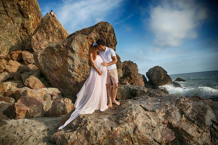 Foto de una pareja embarazada de pie sobre formaciones rocosas junto a la playa.  La mujer lleva un vestido largo rosa y se están abrazando. 