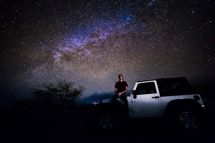 Un fotógrafo de la Vía Láctea sentado en el capó de un coche bajo un cielo lleno de estrellas