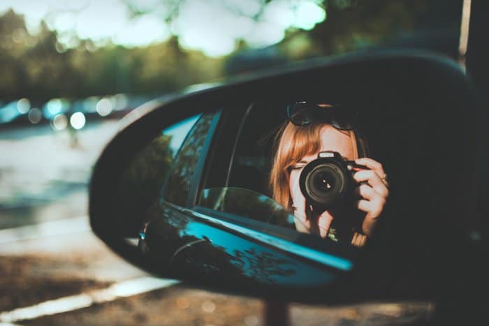 un reflejo de espejo lateral de una mujer usando una DSLR en el asiento trasero de un auto